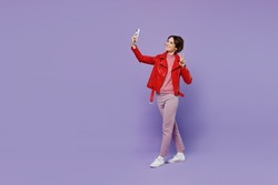 Full body young smiling happy woman 20s wear red leather jacket doing selfie shot on mobile cell phone post photo on social network isolated on plain pastel light purple background studio portrait.