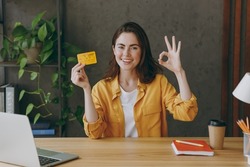 Young successful employee business woman 20s wear casual yellow shirt hold in hand mock up of credit bank card show ok gesture sit work at wooden office desk with pc laptop. Achievement career concept