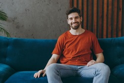 Young smiling cheerful happy fun european man wears red t-shirt sit on blue sofa couch stay at home hotel flat rest relax spend free spare time in living room indoors grey wall. People lounge concept