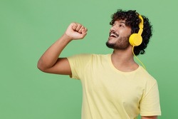 Young singer happy Indian man 20s in basic yellow t-shirt headphones listen to music sing song in microphone isolated on plain pastel light green background studio portrait People lifestyle concept