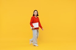 Full size body length fancy smiling happy young woman of Asian ethnicity 20s years old in casual clothes hold under hand laptop pc computer go move isolated on plain yellow background studio portrait