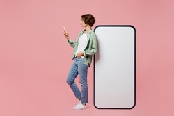 Full body side view young woman wear green shirt white t-shirt near big huge blank screen mobile cell phone smartphone with mockup use mobile cell phone isolated on plain pastel light pink background