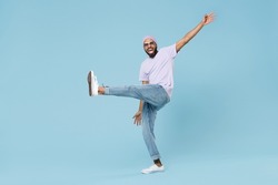 Full body young smiling happy man of African American ethnicity 20s wearing violet t-shirt hat glasses standing with raised up leg outstretched hands isolated on pastel blue background studio portrait