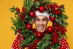 Close up merry fun smiling young man wear red knitted sweater Santa hat posing hold look through Christmas wreath isolated on plain yellow background Happy New Year 2023 celebration holiday concept