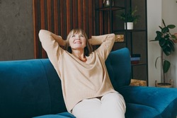 Elderly smiling woman 50s years old wears casual clothes sits on blue sofa hold hands behind neck look overhead dream stay at home flat rest relax spend free spare time in living room indoor grey wall