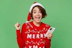 Merry young woman wears knitted xmas sweater Santa hat posing hold in hand use mobile cell phone do winner gesture isolated on plain pastel light green background. Happy New Year 2023 holiday concept
