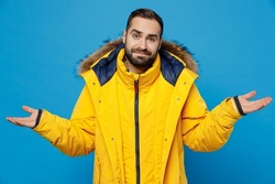 Young caucasian man 20s wear yellow down jacket spread hands shrugging shoulders looking puzzled, have no idea isolated on plain blue color background studio portrait. People winter lifestyle concept