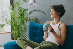 Side view young woman wears white tank shirt drink tea coffee sit on blue sofa couch stay at home hotel flat rest relax spend free spare time in living room indoors grey wall. People lounge concept