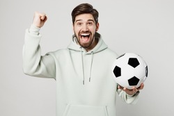 Young excited happy man 20s fan wearing mint hoody do winner gesture cheer up support football sport team hold in hand soccer ball watch tv live stream isolated on plain solid white background studio
