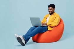Full body smiling happy young man of African American ethnicity he wear yellow shirt sit in bag chair hold use work on laptop pc computer isolated on plain pastel light blue background studio portrait