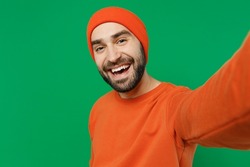 Close up young smiling happy caucasian man 20s wear orange sweatshirt hat doing selfie shot pov on mobile phone look camera isolated on plain green background studio portrait. People lifestyle concept