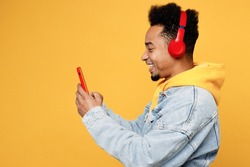 Side view young happy man of African American ethnicity wears denim jacket hoody headphones listen to music use mobile cell phone isolated on plain yellow background studio. People lifestyle concept