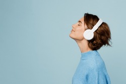 Young happy fun caucasian woman wear knitted sweater headphones listen to music with closed eyes have fun isolated on plain pastel light blue cyan background studio portrait. People lifestyle concept