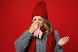 Young sad woman wearing grey sweater scarf hat hold napkin tissue blowing nose sneeze isolated on plain red background studio portrait Healthy lifestyle ill sick disease treatment cold season concept
