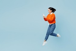 Full body young woman of Asian ethnicity wear orange sweater glasses jump high hold in hand use mobile cell phone isolated on plain pastel light blue cyan background studio. People lifestyle concept