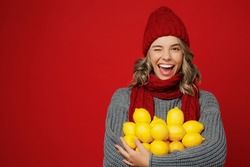 Young happy fun woman wear grey sweater scarf hat hold in hands bunch of lemons wink isolated on plain red background studio portrait. Healthy lifestyle ill sick disease treatment cold season concept