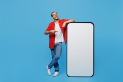 Full body young fun man of African American ethnicity wear red shirt point index finger on big huge blank screen mobile cell phone with mockup area isolated on plain pastel light blue cyan background