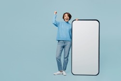 Full body young fun woman wear knitted sweater near big huge blank screen mobile cell phone smartphone with workspace mockup area do winner gesture isolated on plain pastel light blue cyan background