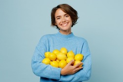 Young smiling cool positive healthy caucasian woman wear knitted sweater look camera hold pile of lemons isolated on plain pastel light blue cyan background studio portrait. People lifestyle concept