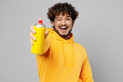 Young cheerful Indian man 20s he wearing casual yellow hoody hold in hand graffiti spray paint bottle point camera on you isolated on plain grey background studio portrait. People lifestyle portrait