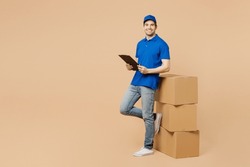 Full body delivery guy employee man wears blue cap t-shirt uniform workwear work as dealer courier stand near stack cardboard boxes hold clipboard papers documents isolated on plain beige background