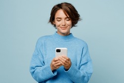 Young smiling happy fun caucasian woman wear knitted sweater hold in hand use mobile cell phone chatting isolated on plain pastel light blue cyan background studio portrait. People lifestyle concept