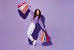 Full body young woman wear pullover hold paper package bags after shopping stand on toes lean back spread hands dance isolated on plain pastel light purple background Black Friday sale buy day concept