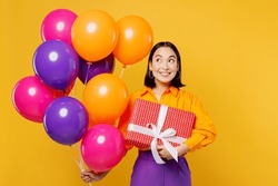 Happy fun smiling amazed young woman wear casual clothes celebrating look at balloons holding present box with gift ribbon bow isolated on plain yellow background. Birthday 8 14 holiday party concept