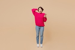 Full body young sad confused puzzled woman wearing pink sweater holdhead using mobile cell phone look camera isolated on plain pastel light beige background studio portrait. People lifestyle concept