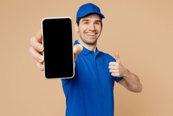 Delivery guy employee man wearing blue cap t-shirt uniform workwear work as dealer courier hold in hand use mobile cell phone with blank screen area show thumb up isolated on plain beige background