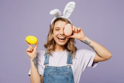 Young smiing cheerful fun woman wearing casual clothes bunny rabbit ears cover eye with colorful eggs isolated on plain pastel light purple background studio portrait. Lifestyle Happy Easter concept