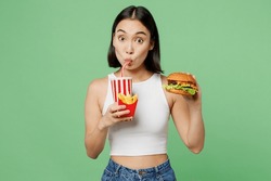 Young shocked fun woman wears white clothes hold bottle of soda pop cola eat burger french fries isolated on plain pastel light green background. Proper nutrition fast food unhealthy choice concept