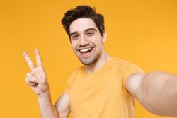 Close up young smiling unshaved friendly caucasian man 20s wear basic blank print design t-shirt doing selfie shot on mobile phone show victory sign isolated on yellow color background studio portrait