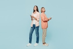 Full body fun happy elder parent mom with young adult daughter two women together wear casual clothes hold in hand mobile cell phone look overhead isolated on plain blue background. Family day concept