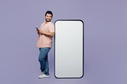 Full body side view fun young Indian man he wear pink shirt white t-shirt casual clothes big huge blank screen mobile cell phone with area using smartphone isolated on plain pastel purple background