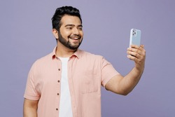 Young Indian man he wear pink shirt white t-shirt casual clothes doing selfie shot on mobile cell phone post photo on social network isolated on plain pastel light purple background. Lifestyle concept