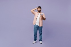 Full body sad shocked scared young Indian man he wear pink shirt white t-shirt casual clothes use mobile cell phone hold head isolated on plain pastel light purple background studio. Lifestyle concept