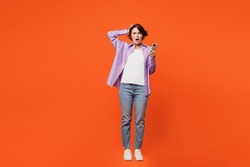 Full body angry mad astonished sad young woman she wear purple shirt white t-shirt casual clothes hold head use mobile cell phone isolated on plain orange background studio portrait. Lifestyle concept