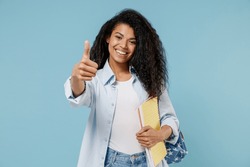Young woman of African American ethnicity student wear denim clothes backpack hold books show thumb up gesture isolated on blue color background studio. Education in high school university concept.