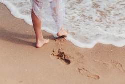 Cropped photo of feet of  bride in white lace wedding dress on clean beach. Female legs on sandy beach and  waves of sea or ocean, summer vacation at water. Wedding, rest, relax concept.