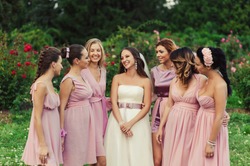 Beautiful happy smiling bride gossip with bridesmaids in light trendy pink dresses on walk outdoors in green summer floral blossom park. Friends, maid of honor, female friendship, wedding concept.