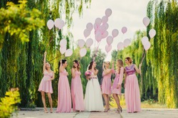 Beautiful happy smiling bride with bridesmaids in light trendy pink dresses on walk outdoors holding pink balloons in green summer city park. Friends, maid of honor, female friendship, wedding concept