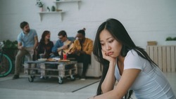 Young asian girl feels upset and isolated while her flatmates celebrating party at home