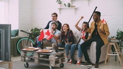 Multi ethnic group of friends sports fans with Canadian national flags watching hockey championship on TV together cheering up their favourite team at home indoors