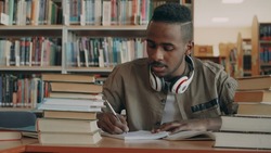 african american young handsome guy with big headphones is sitting at table with books and writing in his copybook