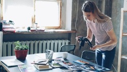 Young attractive female blogger using digital camera to shoot colourful pictures from travel displayed on table. She is moving photos and making different collages. Loft style office in background.