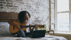 Curly african american teenager girl learning to play guitar using laptop computer sitting on bed at home