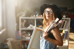 Pretty caucasian long-hair female artist in cute black hat and eyeglasses holding brush and canvas in light contemporary studio