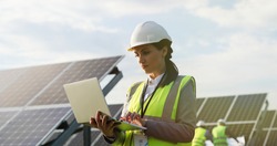Portrait of electrician engineer in safety helmet and uniform using laptop checking solar panels. Female technician at solar station.