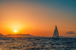 One sailing boat floating on the blue Aegean Sea during sunrise / sunset with open main sail and genoa. Orange sky and sun with islands in background for copy space